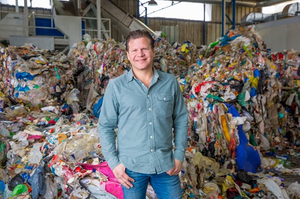 A man in a blue collared shirt and blue jeans smiling and standing inside a recycling plant in front of piles of materials to be recycled