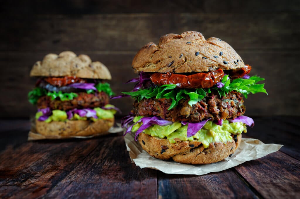Two veggie burgers on sprouted bunes with lettuce, tomatoes, cabbage and guacamole placed on a dark brown wooden table