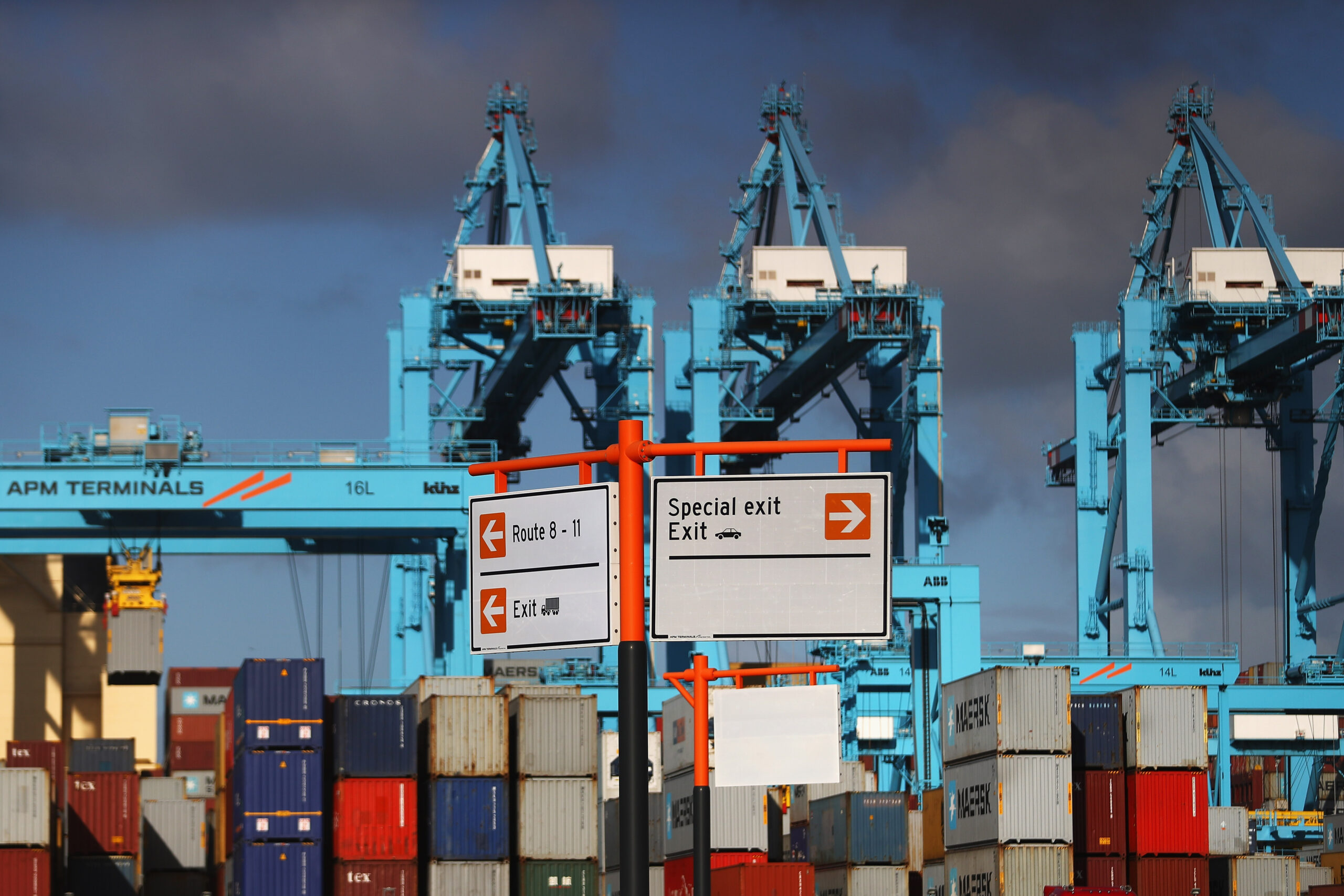 General view of Rotterdam Port that includes multi-colored shipping containers and blue cranes. Two signs hang off orange boles indicating directions.