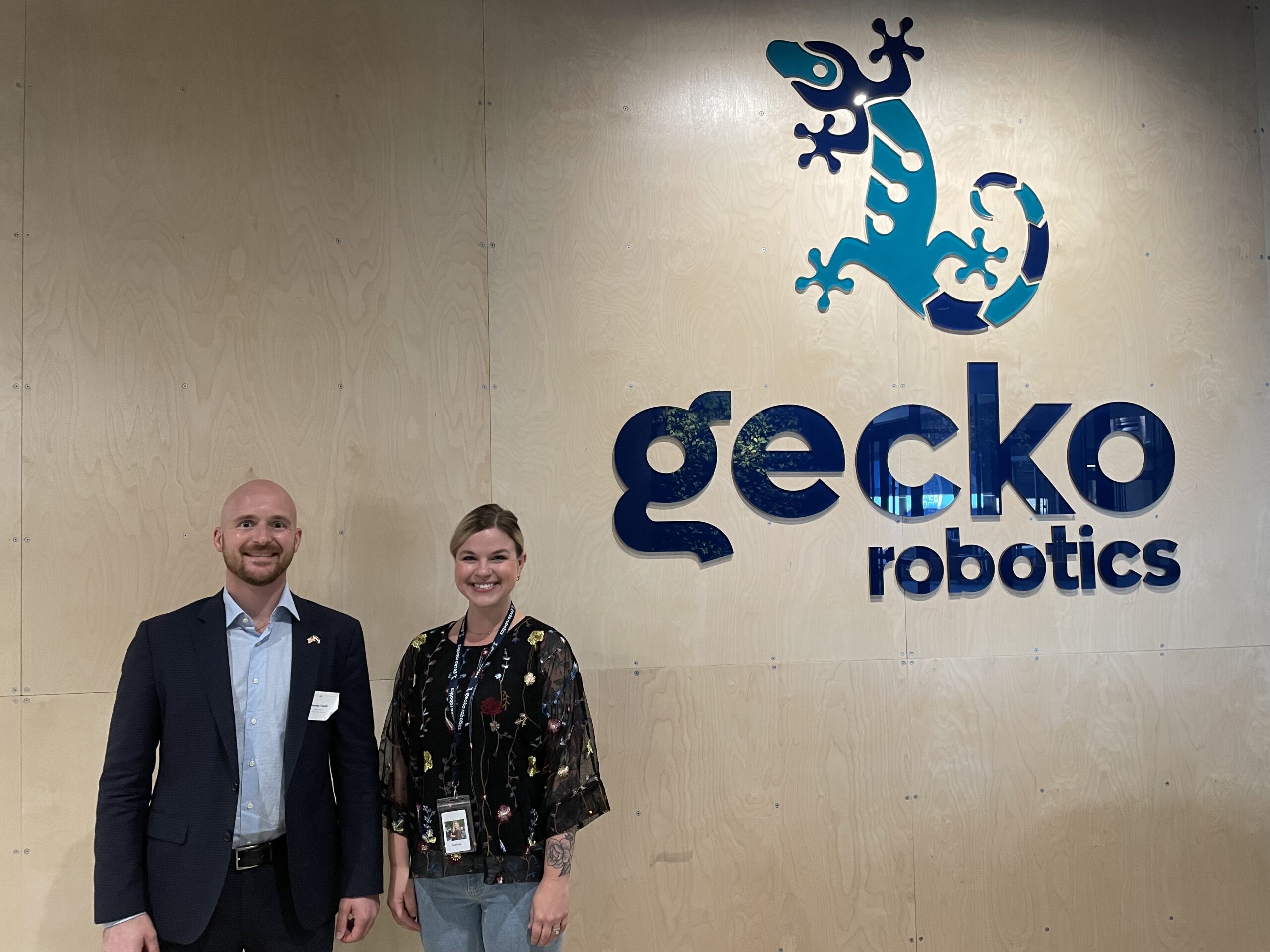 A man and woman smile at the camera, the words, "Gecko Robotics" and a blue gecko emblem on a wall behind them — Invest in Holland.