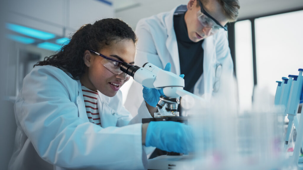 Two young scientists — a man and a woman in white lab coats — use a microscope and digital tablet.